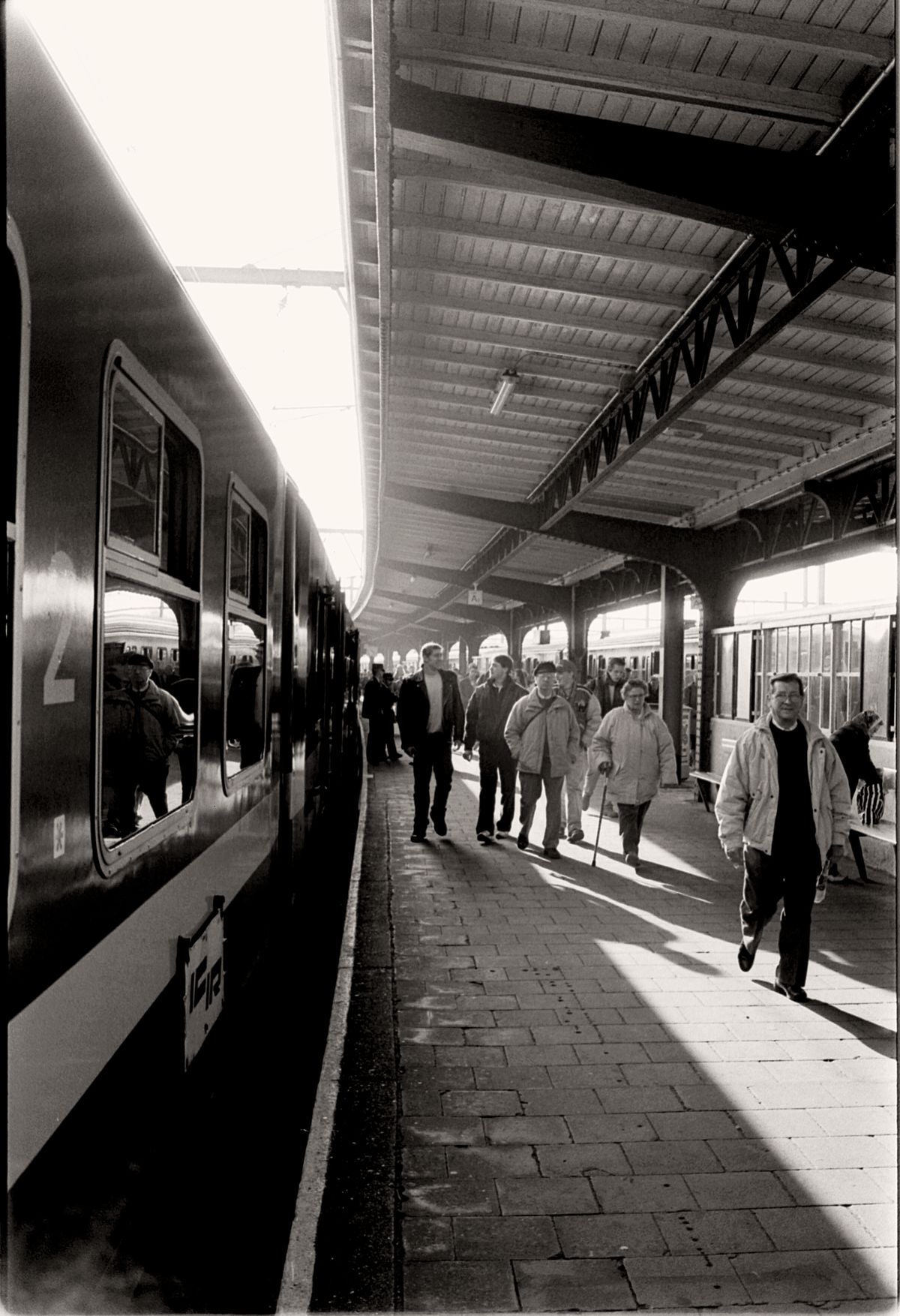 Black-and-white photo of passengers walking along a sunlit train station platform, one leaning on a cane
