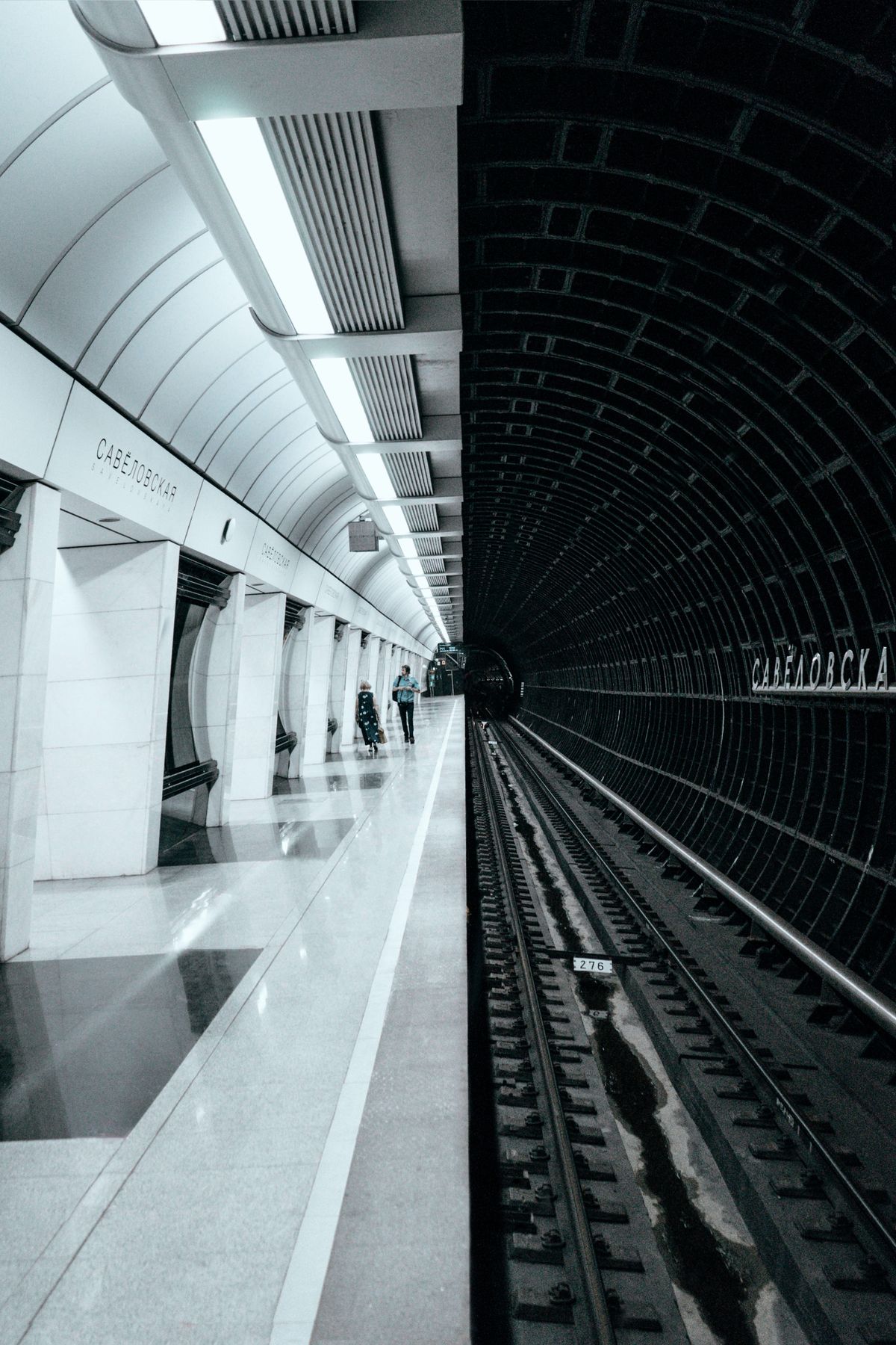 Wide-angle monochrome photo of a modern subway platform and tunnel with two commuters walking toward the train
