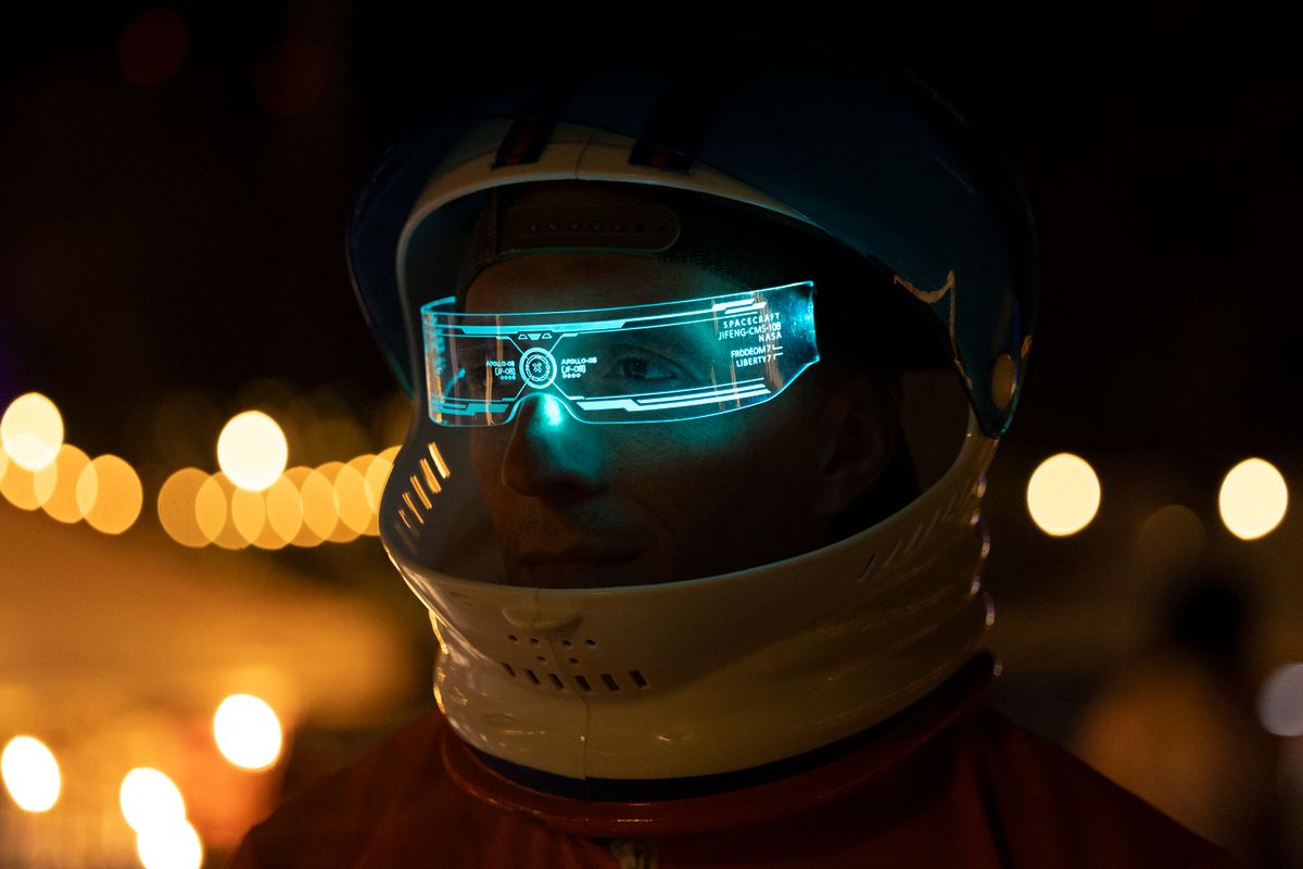Close-up of a futuristic helmet visor with a glowing cyan holographic HUD display, dark background with warm bokeh lights