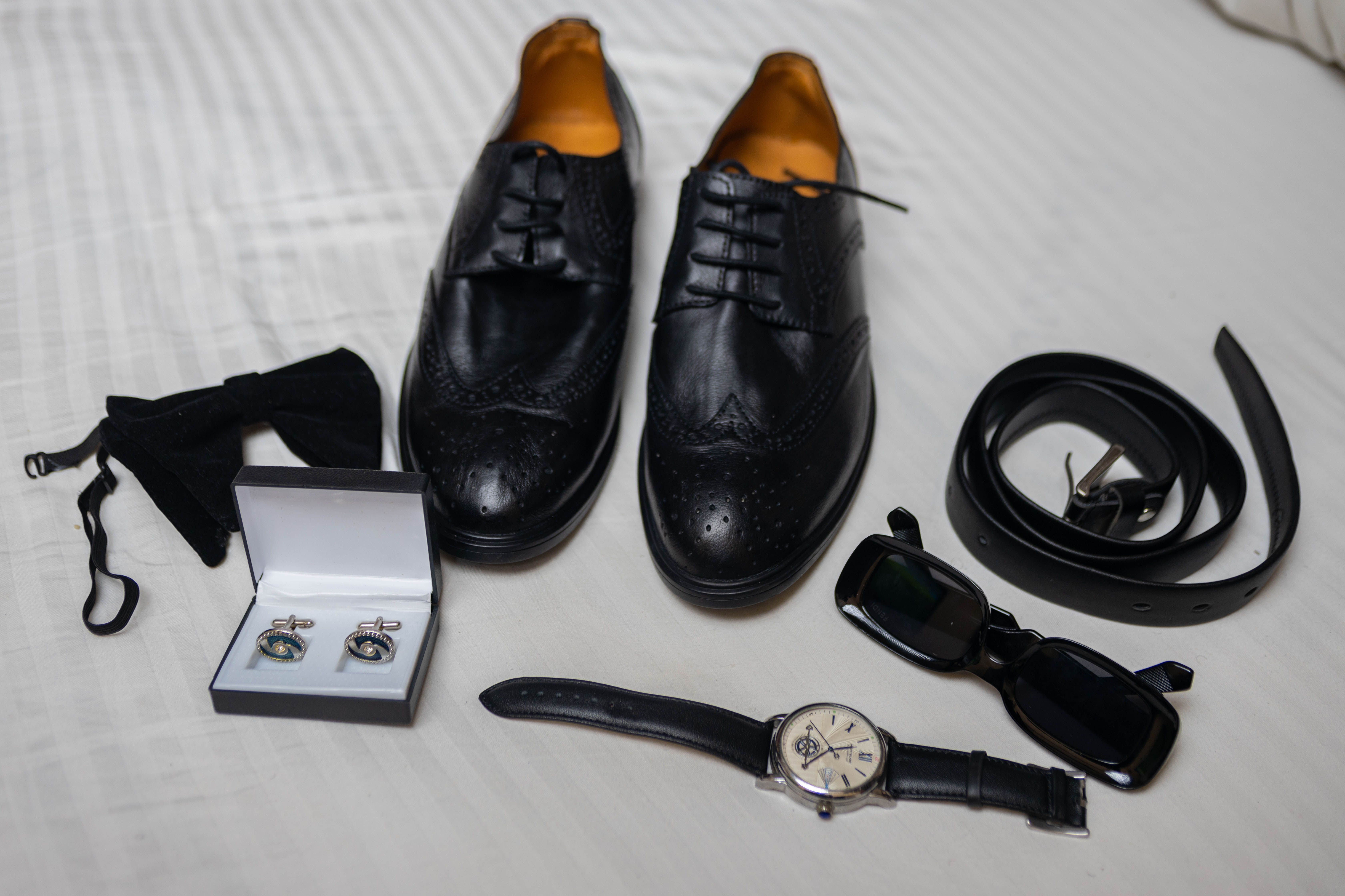 Black leather brogues, belt, sunglasses, bow tie, cufflinks, and wristwatch arranged as a flat lay on a light textured sheet