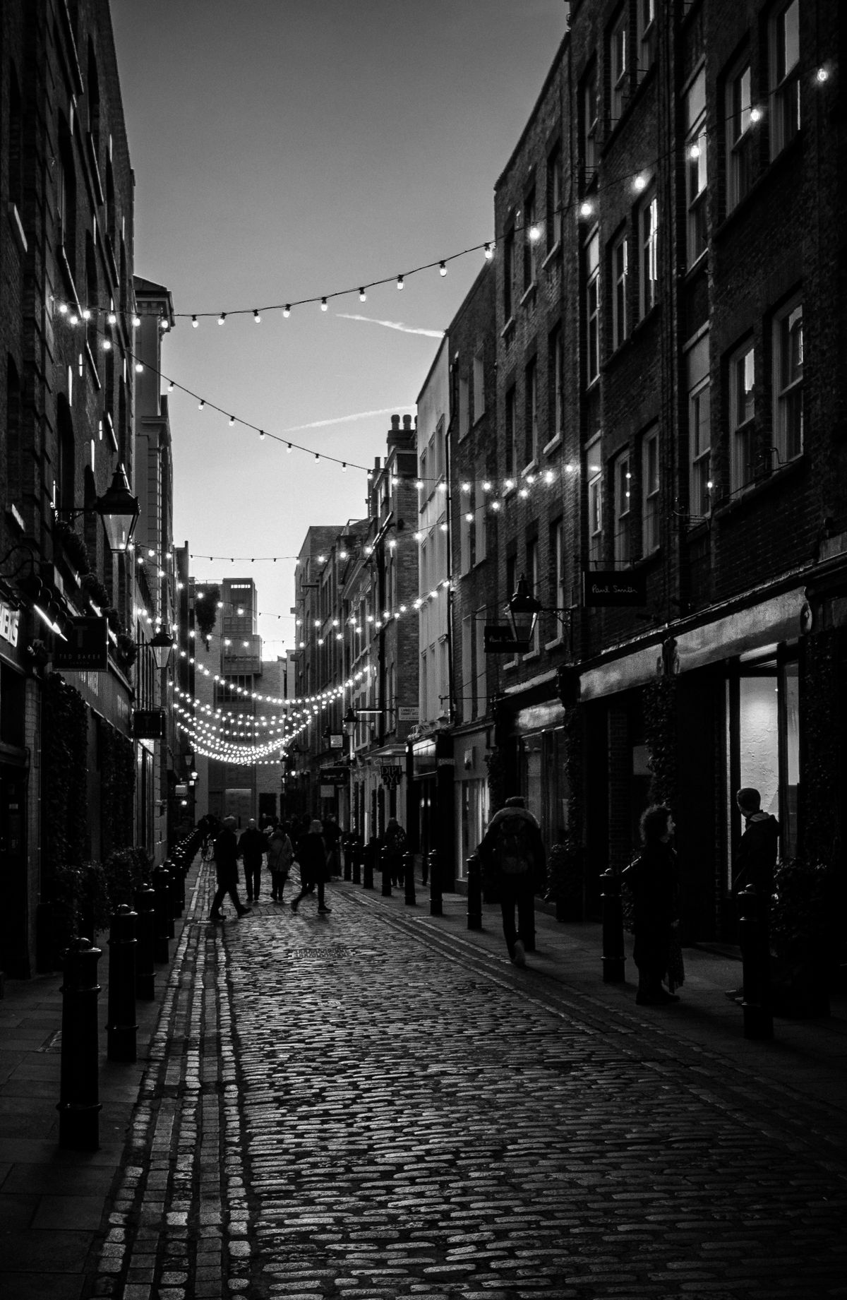 Black-and-white photo of pedestrians walking down a cobblestone lane strung with string lights at dusk