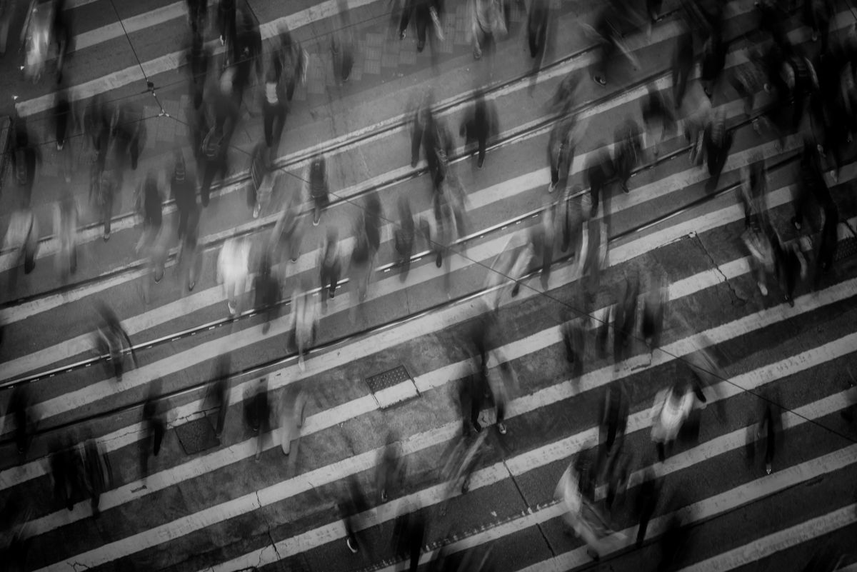 Overhead long-exposure black-and-white photo of pedestrians crossing a striped crosswalk, blurred in motion