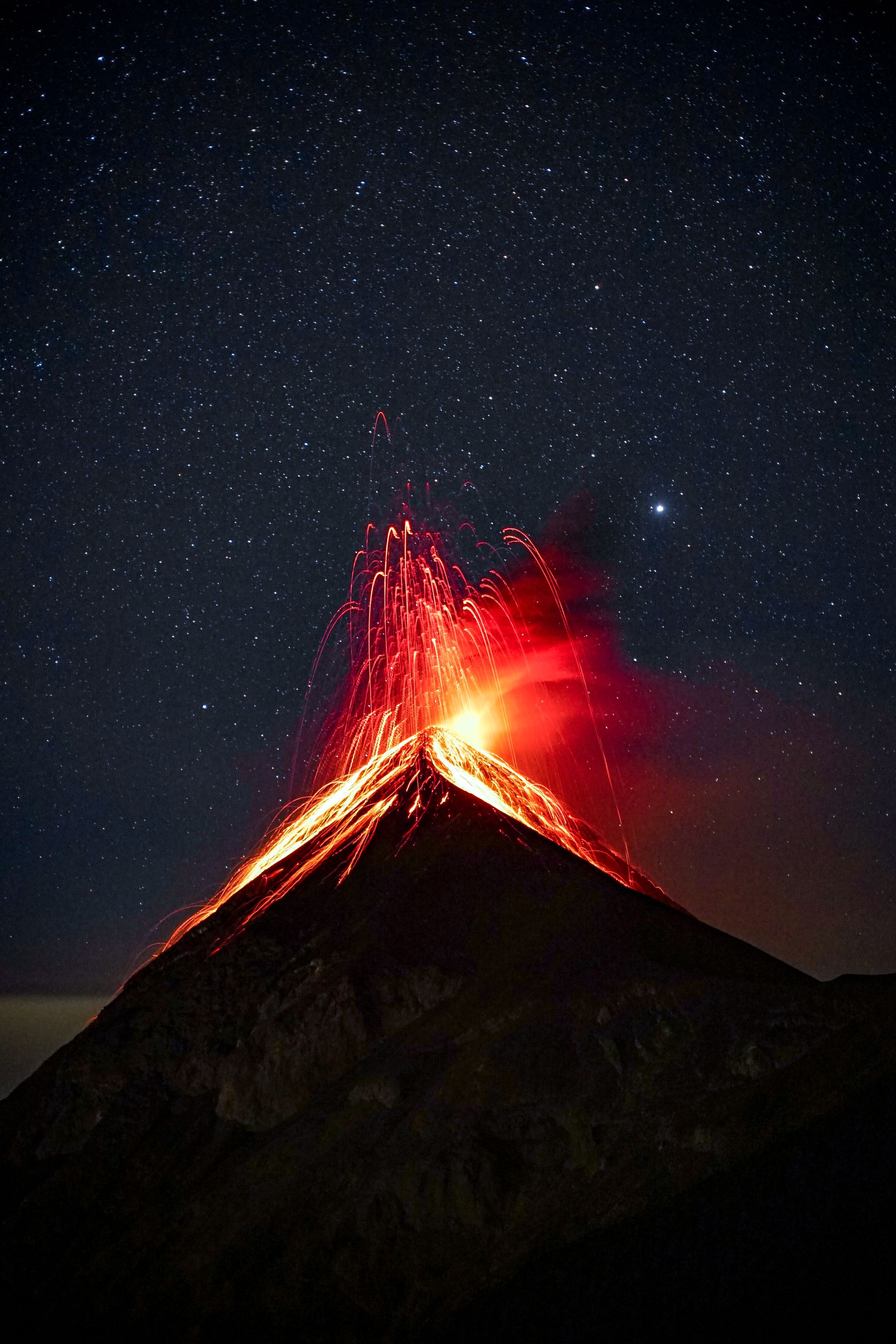 Volcán de Fuego erupting at night, glowing lava fountains arcing over the silhouetted cone under a dense, star-filled sky