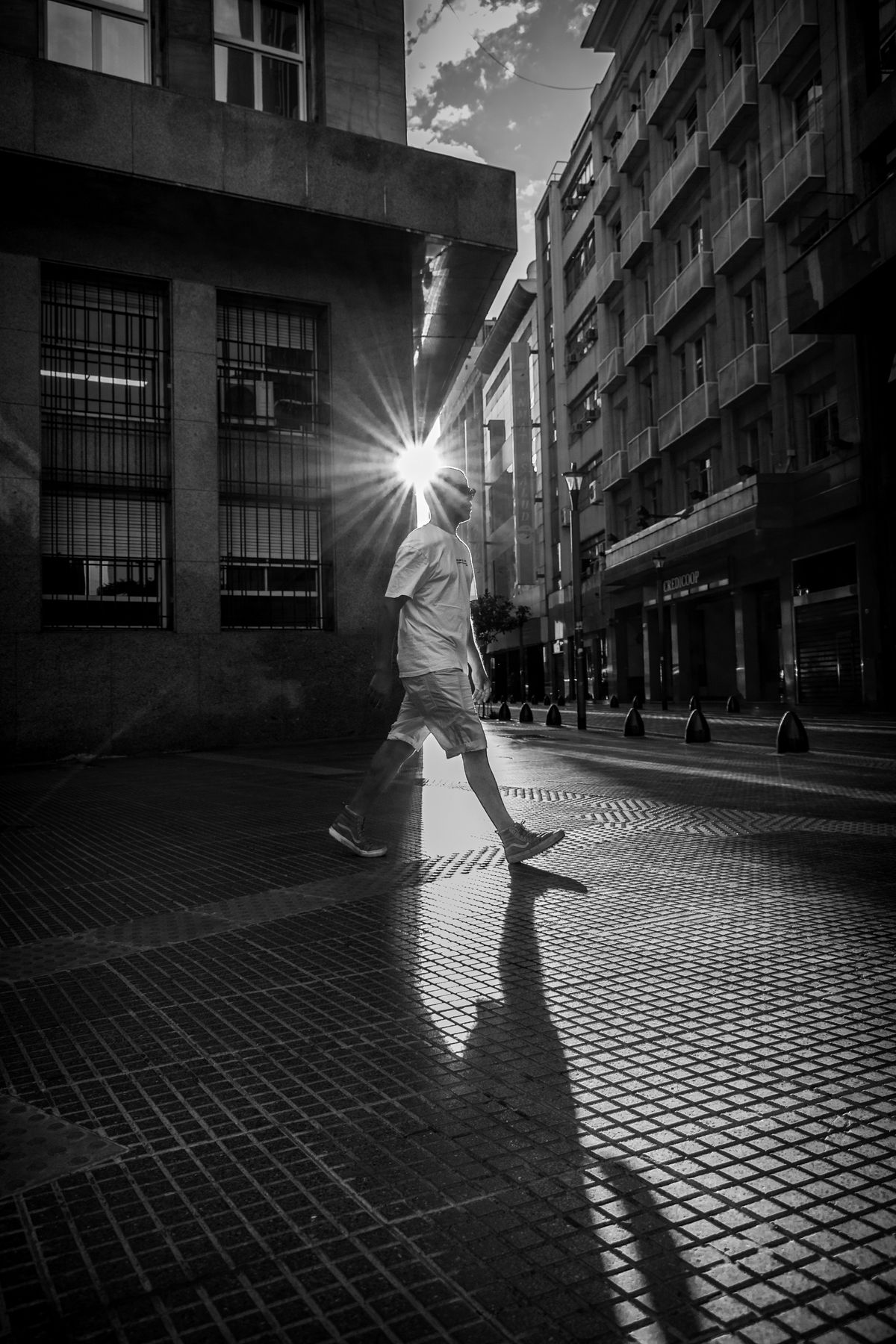 High-contrast black-and-white street photo of a man walking past a sunburst between city buildings