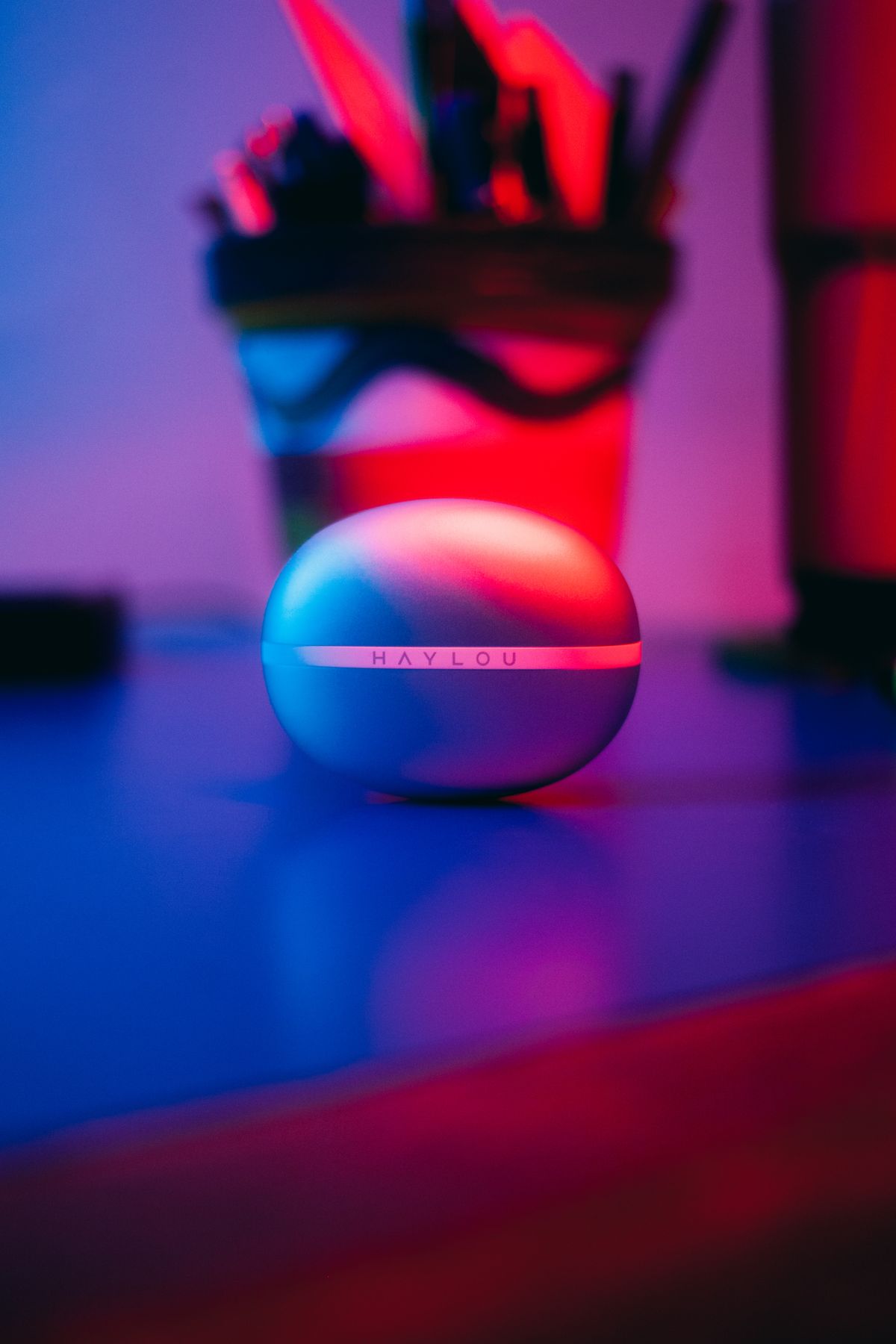 Matte white wireless earbuds case lit by red and blue neon gel lighting on a reflective dark desk