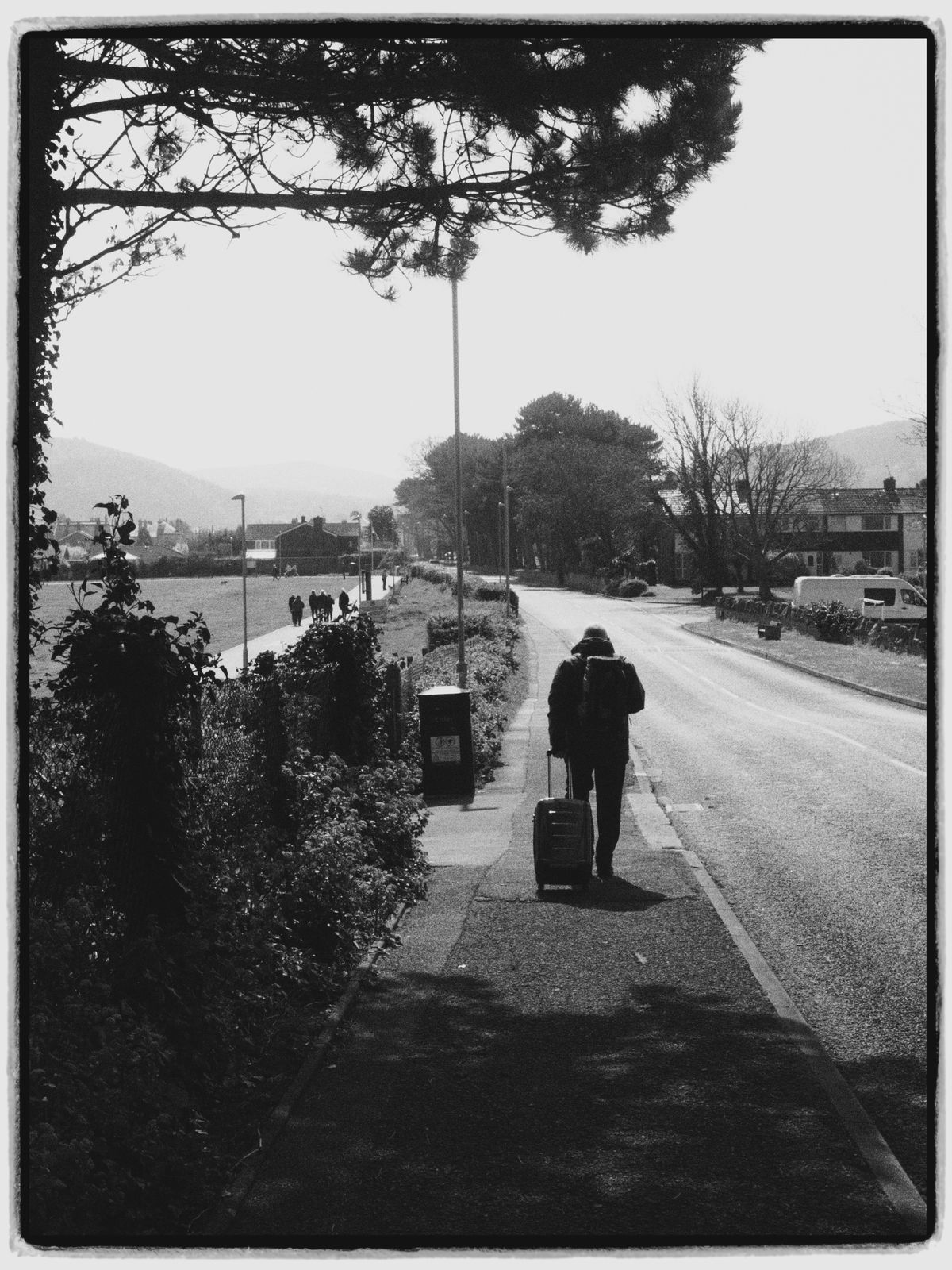 Black-and-white photo of a lone traveler pulling a suitcase down a quiet backlit suburban street