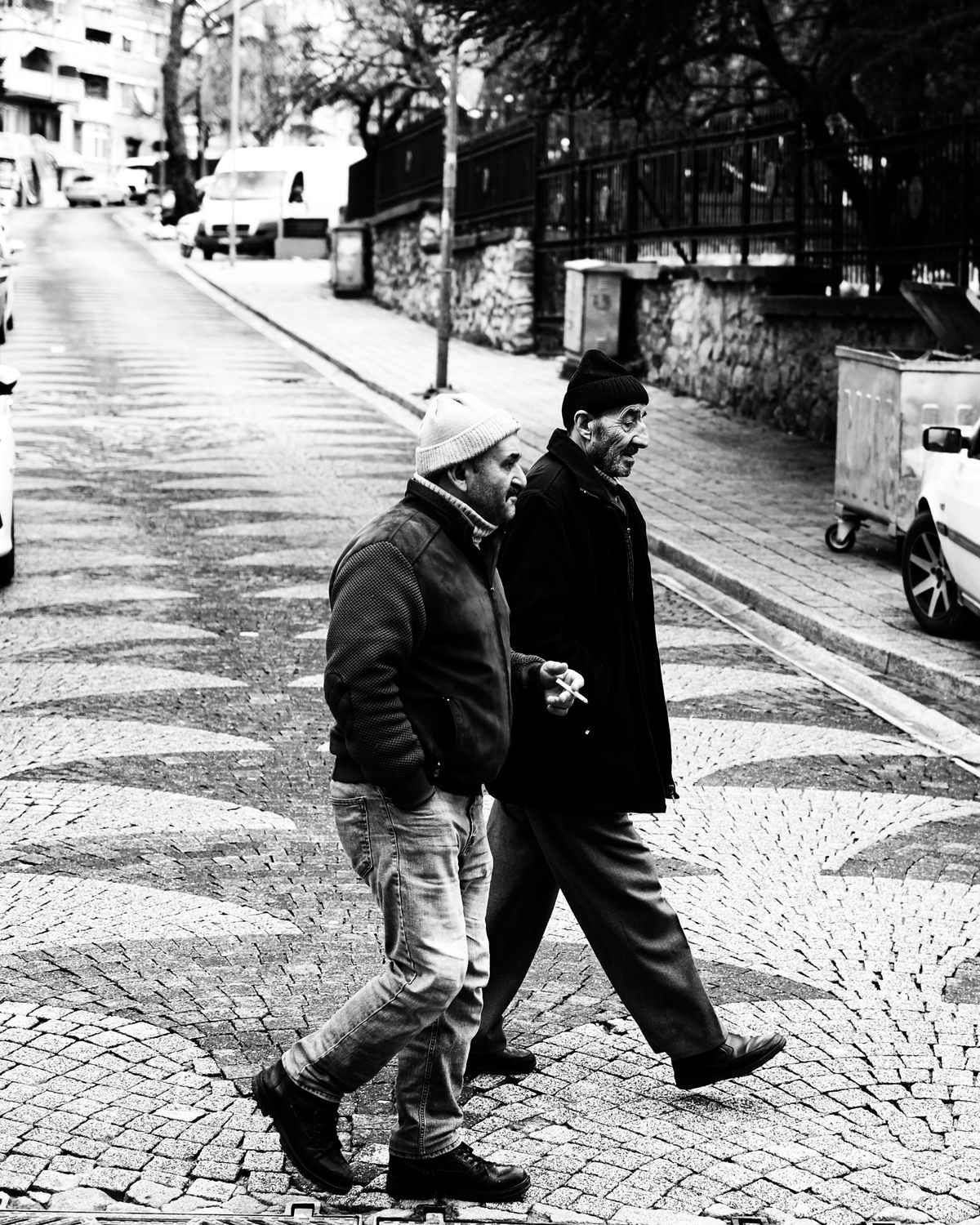 Black-and-white candid photo of two older men walking and talking on a cobblestone street