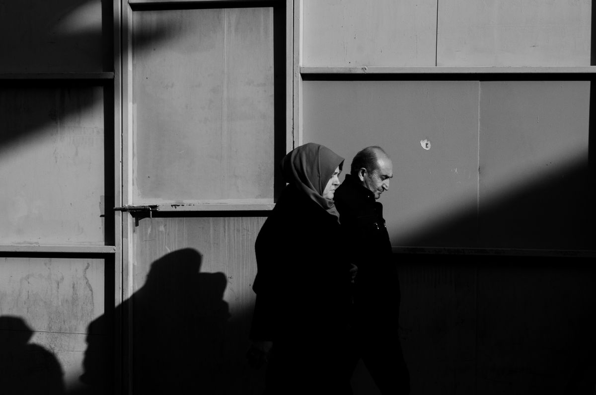 Black-and-white profile of two people walking close together past a sunlit wall, casting long shadows